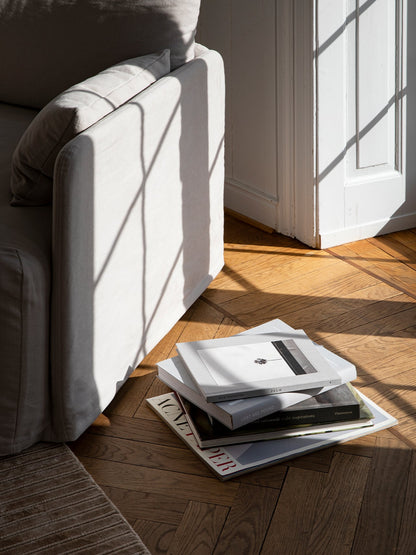Sunlight casts a window shadow on a light grey sofa. A stack of coffee table books rests on the herringbone parquet floor.