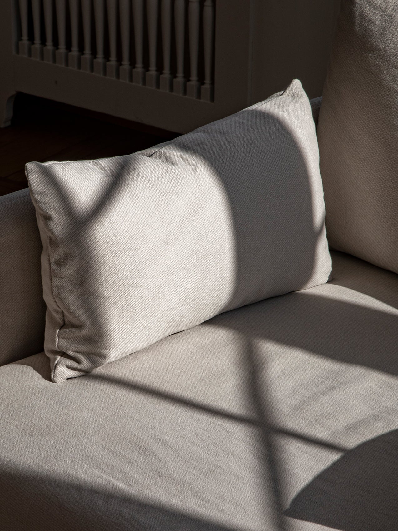 A close-up of a light grey textured fabric cushion resting on a sofa, with sunlight casting an arched shadow across it.
