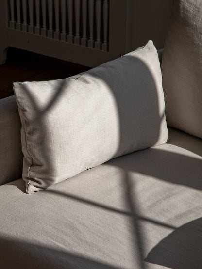 A close-up of a light grey textured fabric cushion resting on a sofa, with sunlight casting an arched shadow across it.