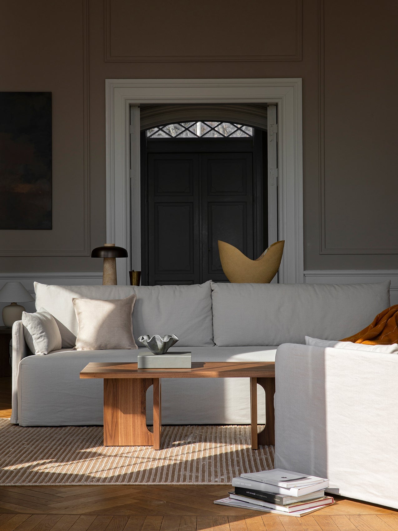 A light grey Eave Modular Sofa in a living room with a wooden Passage Lounge Table. Sunlight streams across a striped rug.