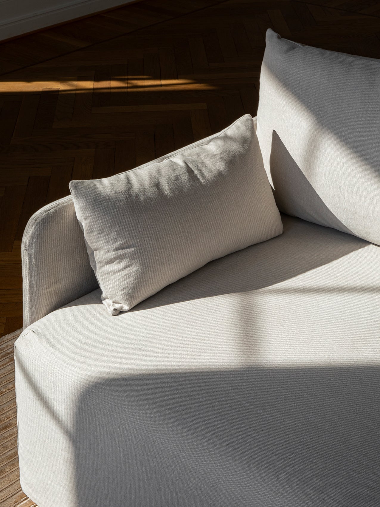 A shot of a light grey upholstered sofa with a plush cushion. Sunlight creates strong shadows across the textured fabric.