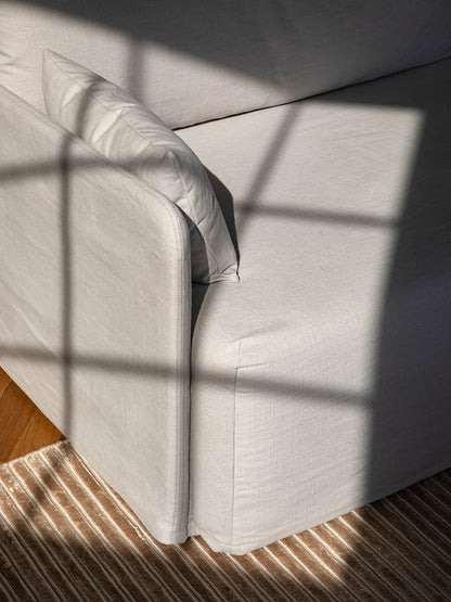 A close-up shot of a light-coloured fabric sofa. Sunlight casts a shadow from a window frame across the armrest and cushion.