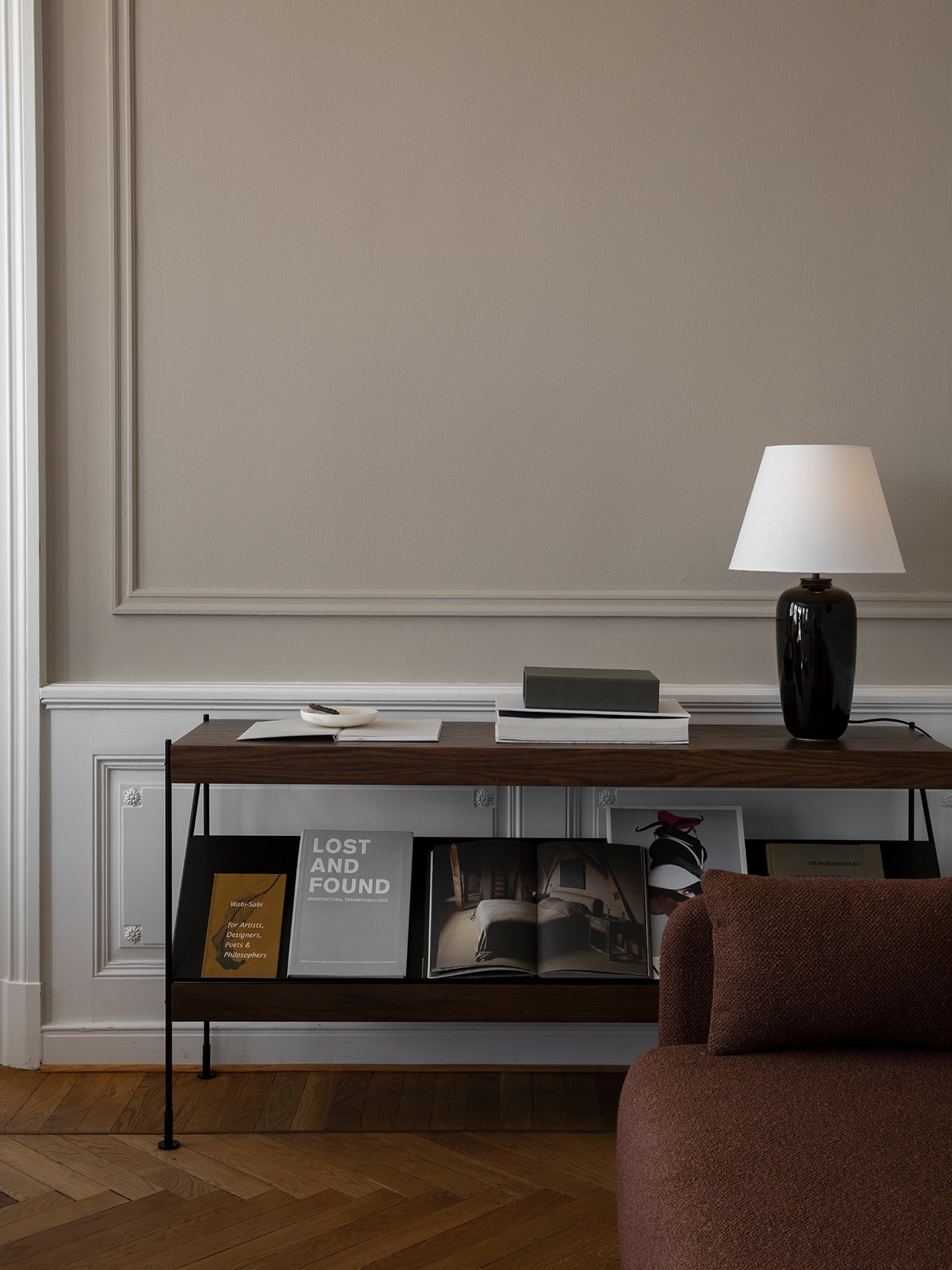 A living room with the Audo Échasse Shelf styled with a black Torso Table Lamp and books, next to a brown Eave Sofa.