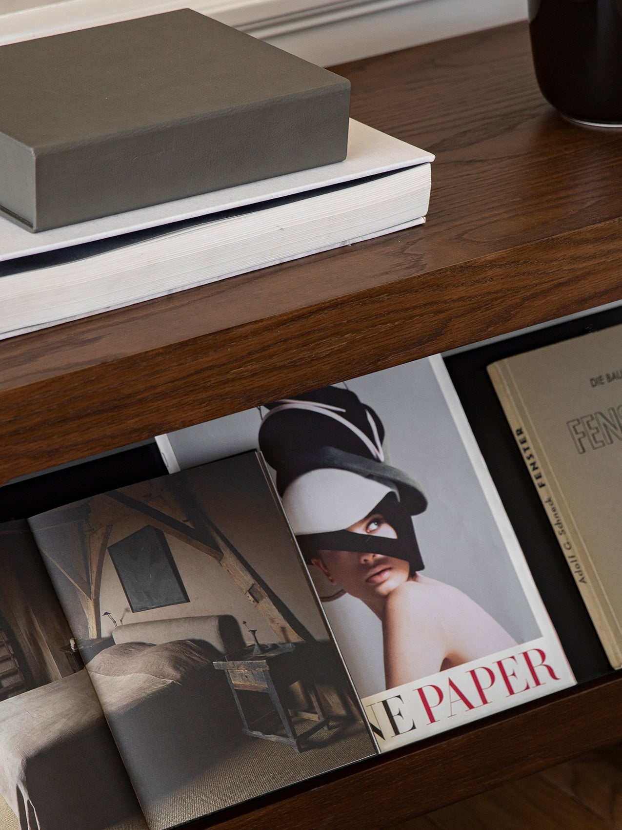 A close-up of a dark wood shelf, with a grey box on a stack of books, and magazines displayed in the open compartment below.