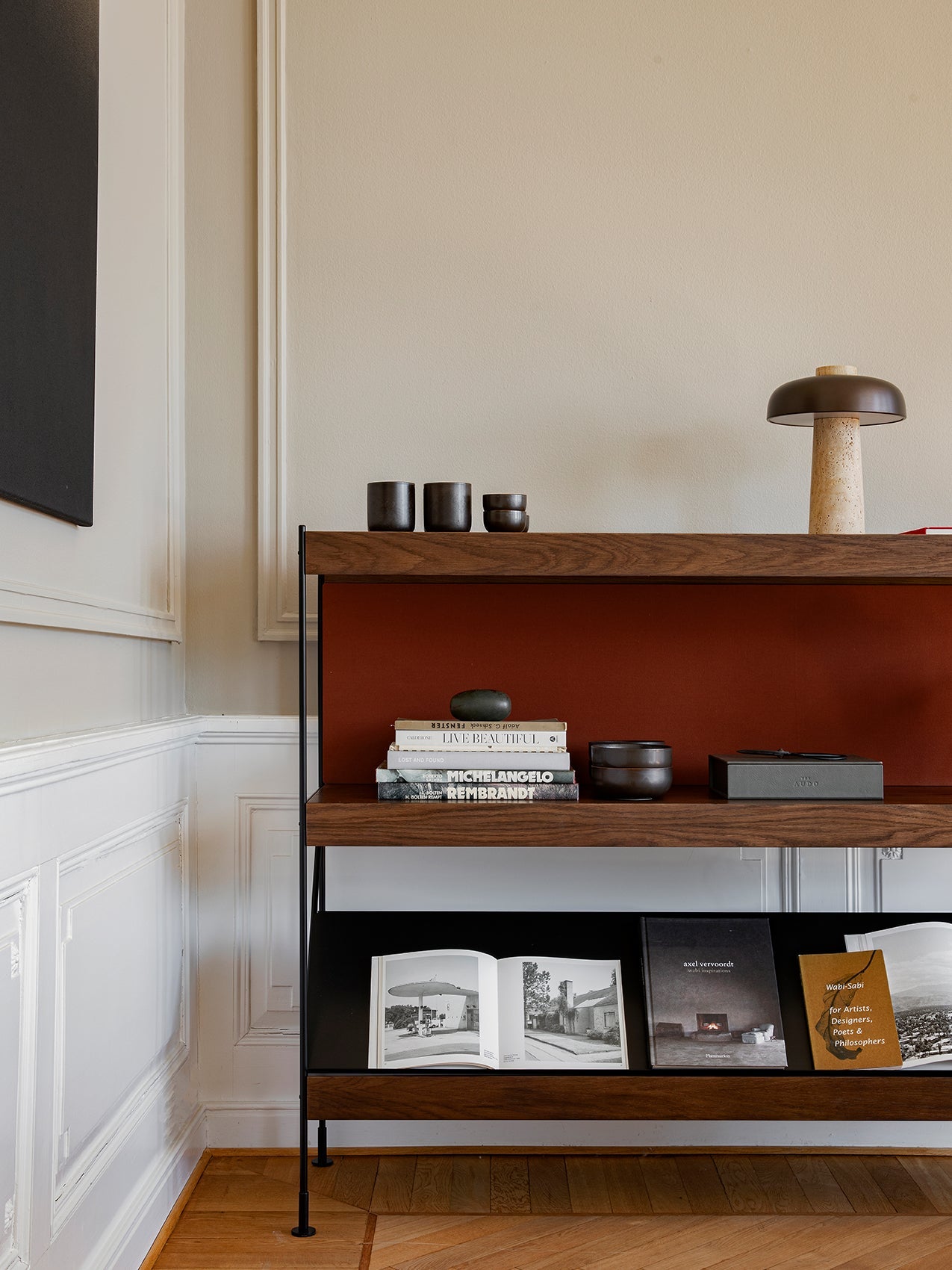 A dark wood shelving unit styled with books, a mushroom lamp, and ceramics, set against a beige wall with white panelling.