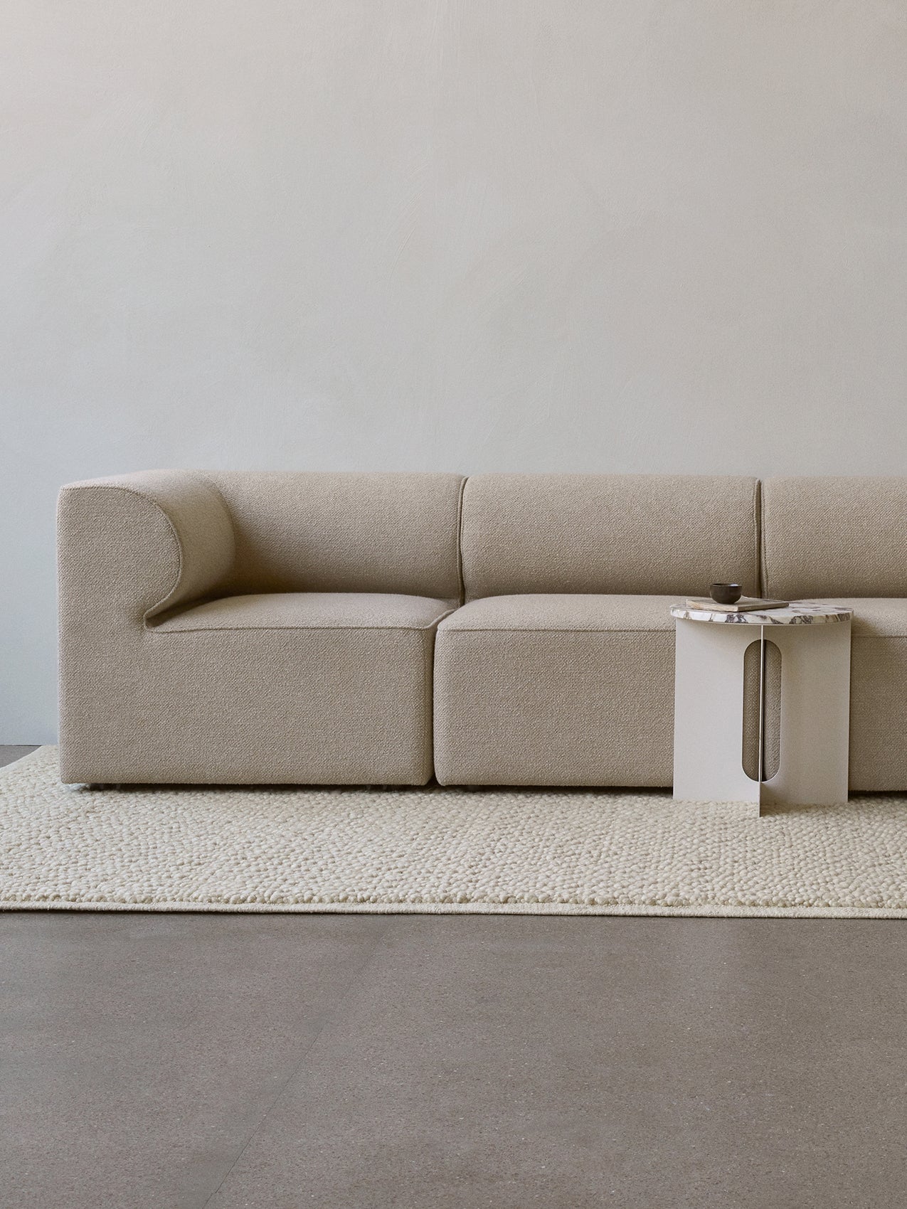 A close-up of the light beige Eave Modular Sofa with its curved armrest, placed on a cream rug next to a marble side table.