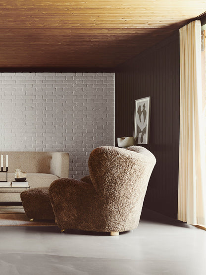 The Tired Man Lounge Chair in Sahara sheepskin, seen from behind in a room with a grey brick wall and dark wood panels.