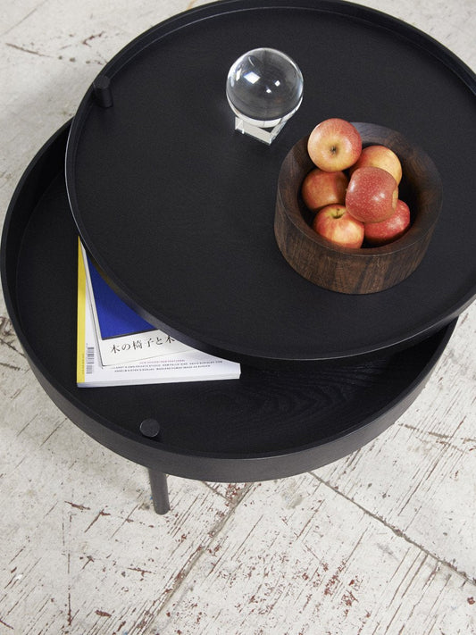 A black Turning Table, its top rotated to show a magazine in storage. On top is a bowl of apples and a glass orb.