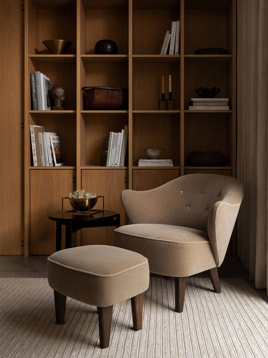 A beige Ingeborg Lounge Chair and Ingeborg Ottoman sit on a striped rug in front of a large wooden shelving unit.