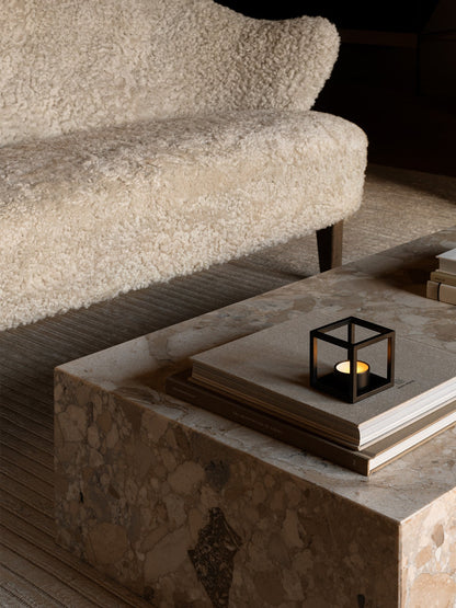 A black Kubus T tealight holder with a lit candle sits on books on a marble table, next to a cream bouclé Ingeborg Sofa.