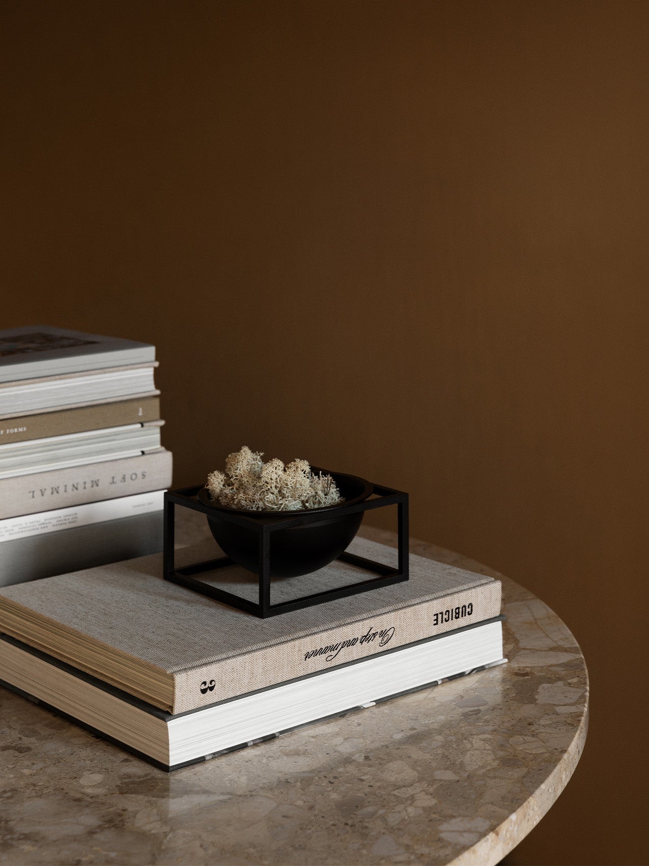 The black by Lassen Kubus Bowl Centerpiece Small, filled with white moss, sits atop a book on a round marble table.