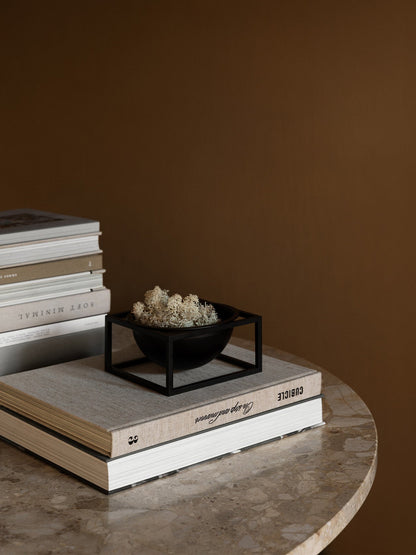 The black by Lassen Kubus Bowl Centerpiece Small, filled with white moss, sits atop a book on a round marble table.
