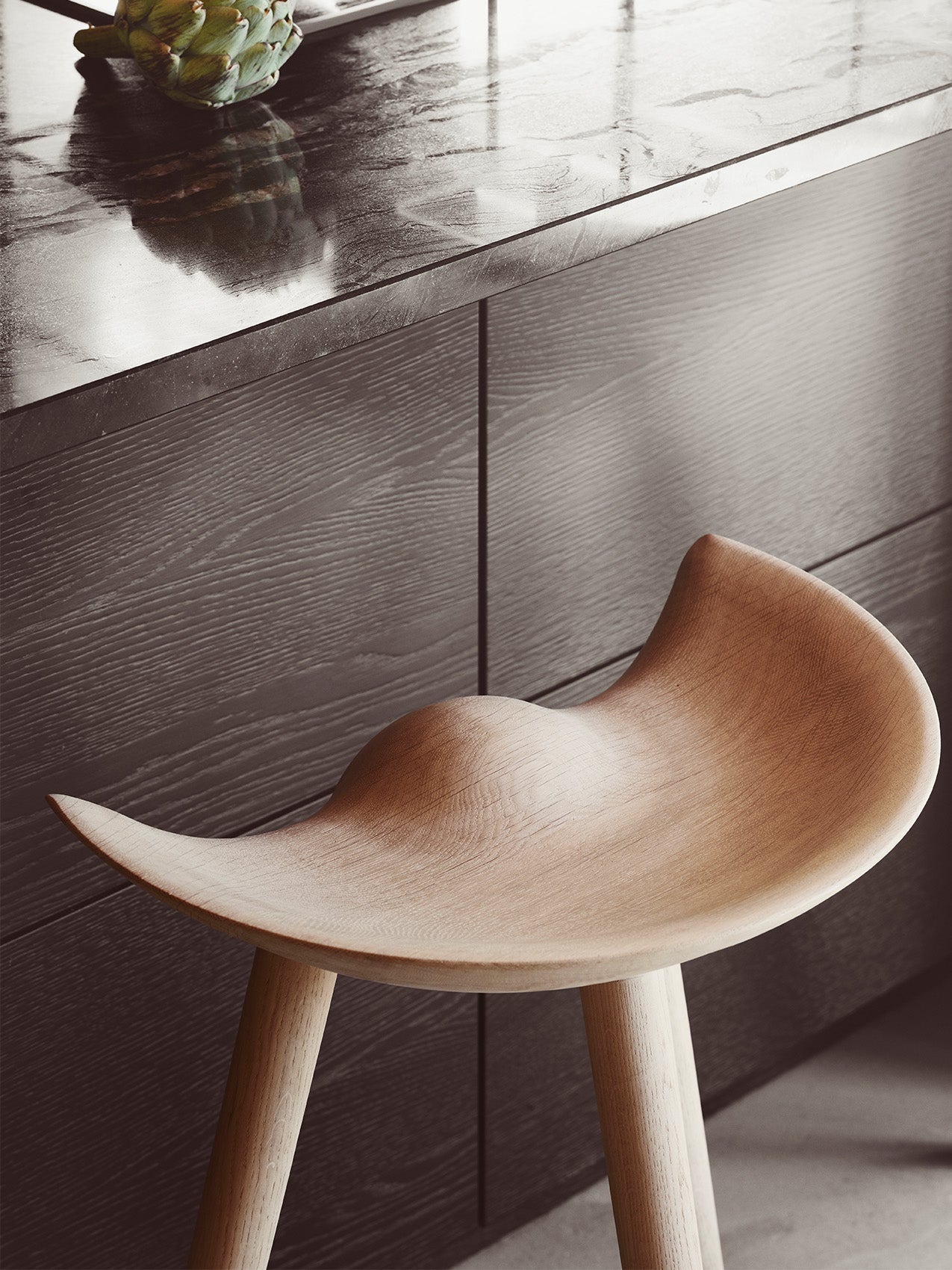 Close-up of the ML 42 Bar Stool in light wood, showcasing the sculpted seat against a dark kitchen counter with a marble top.