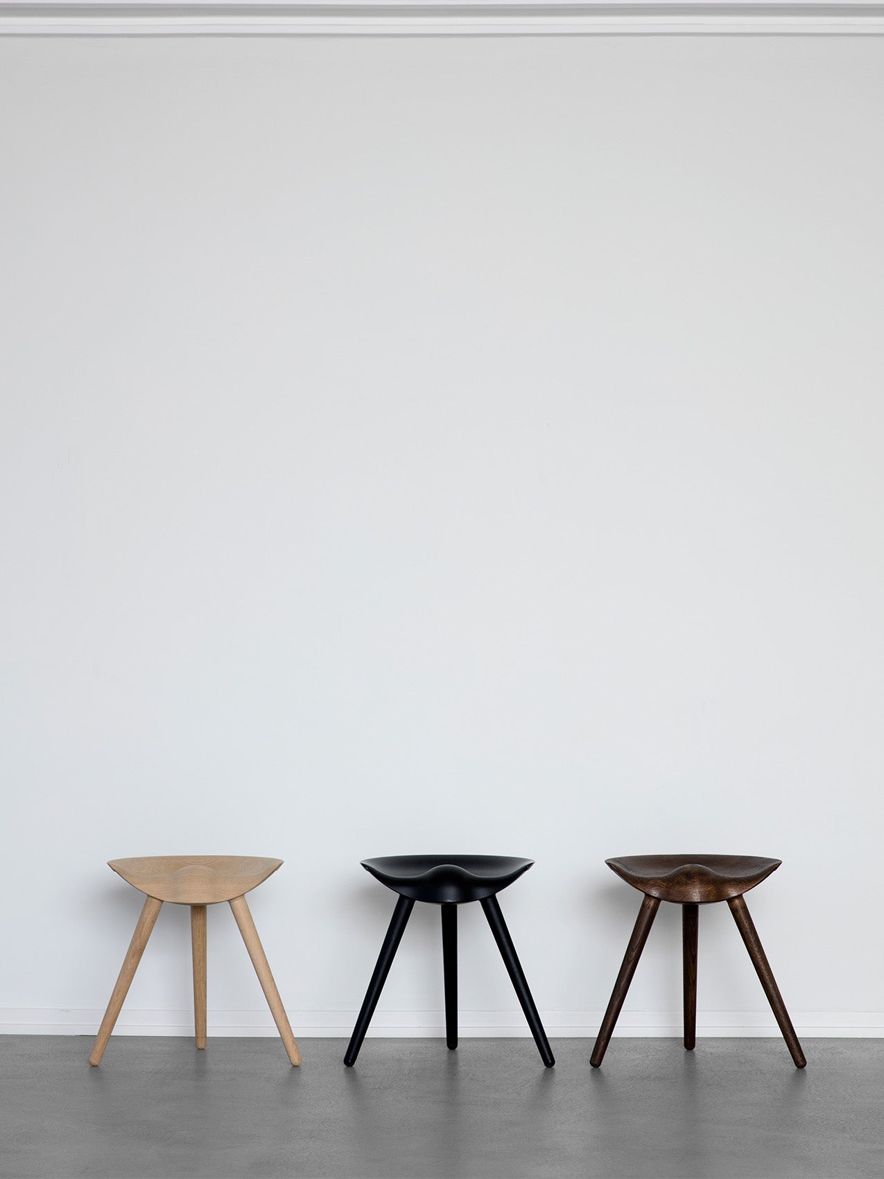 Three sculptural ML 42 Stools in light oak, black, and dark brown wood finishes, displayed in a row against a white wall.