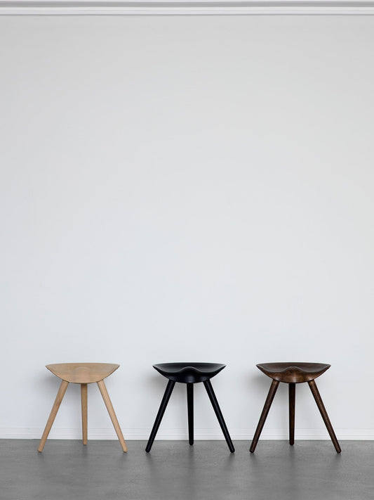 Three sculptural ML 42 Stools in light oak, black, and dark brown wood finishes, displayed in a row against a white wall.
