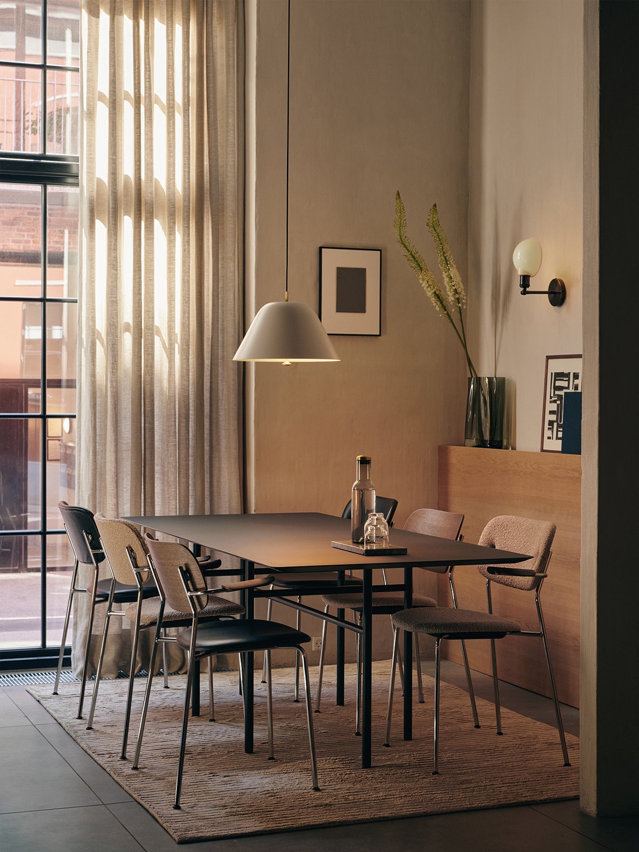 A sunlit dining room featuring several Co Dining Chairs with armrests in various upholsteries around a large black table.
