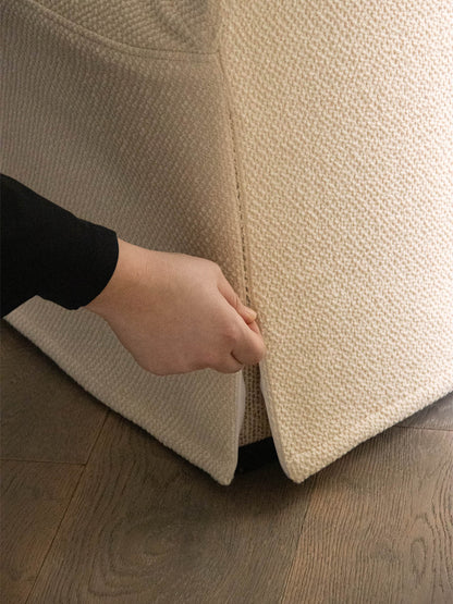A close-up of a hand lifting the textured, cream-coloured Loose Cover for the Tearoom Lounge Chair.